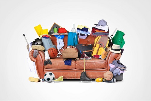 A sofa covered and surrounded by a variety of mixed household items and rubbish, including clothing, sports equipment such as a basketball and soccer ball, shoes, a hat, a backpack, and small miscellaneous objects like a toy car, a yellow plastic container, and a cardboard box. The items appear to be haphazardly piled, occupying the entire seat and extending onto the floor, with some objects leaning against the sofa arms. The background is plain and light, emphasizing the cluttered scene typical of rubbish removal jobs undertaken by Office Clearance Millbank in or near London postcode areas. The materials visible include fabric, plastic, and cardboard, with textures ranging from soft textiles to rigid containers. The scene suggests a cleanout or clearance needing professional rubbish collection, fitting within the broader rubbish removal services offered in the Millbank or Westminster areas.