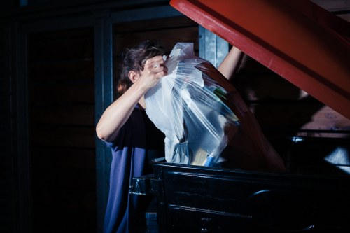 A person wearing a dark T-shirt is seen inside a waste storage area, emptying a large transparent plastic bag filled with various office rubbish, including papers and folders, into a black skip or bin. The environment appears to be an indoor or sheltered space with wooden paneling and metal supports visible in the background. The person is lifting the bag above the bin, with some of the waste already beginning to be deposited inside. The scene is lit by artificial lighting, highlighting the textures of the plastic bag, the waste papers, and the dark finish of the bin, reflecting a typical rubbish removal process. This image relates to professional office clearance services offered by Office Clearance Millbank, providing waste management solutions within the Millbank area for commercial clients.