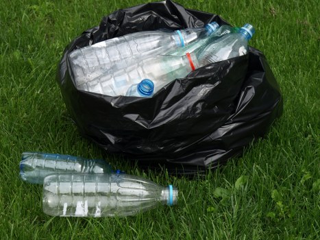A black plastic rubbish bag placed on a grassy surface outdoors, partially filled with various discarded transparent plastic bottles with blue caps and labels. Several bottles are visible both inside the bag and lying on the grass in front of it, indicating collection of recyclable waste. The background features vibrant green grass, suggesting a garden or park setting, with natural daylight illuminating the scene. The image reflects a typical waste collection scenario that a company like Office Clearance Millbank may handle as part of rubbish removal services, emphasizing the collection of plastic waste for disposal or recycling in the context of local environmental efforts in Millbank or nearby London areas.