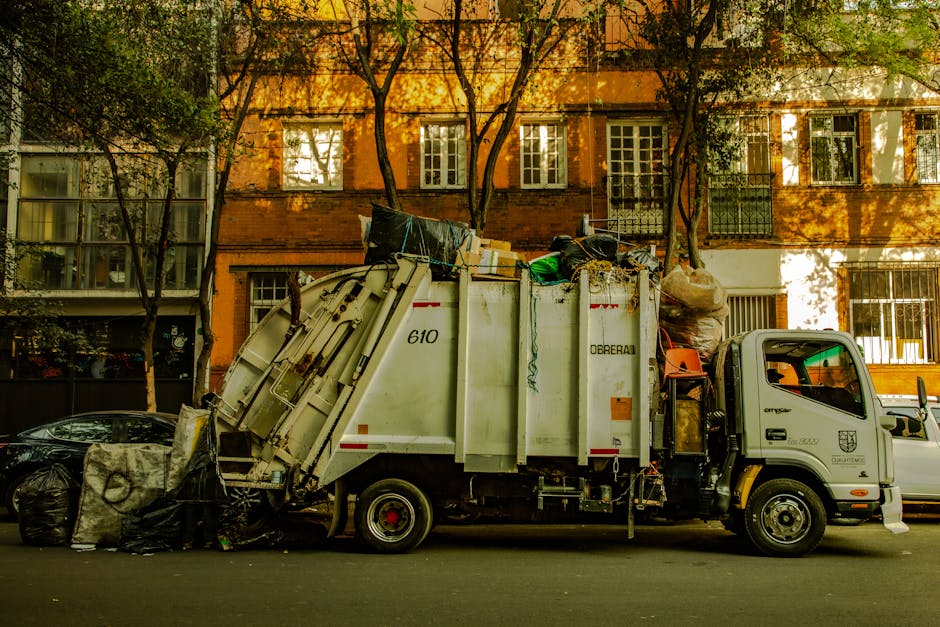 A collection of black trash bags, cardboard boxes, and discarded packaging materials piled in front of a red metal door set into a rough-textured concrete wall. The trash appears to be informal waste,