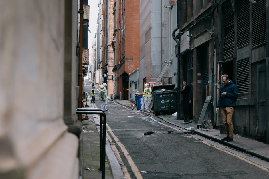 A narrow urban street scene during daytime showing a group of construction workers in high-visibility vests and helmets actively engaged in rubbish removal and site clearance. The workers are position