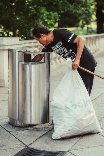 A narrow urban street scene during daytime showing a group of construction workers in high-visibility vests and helmets actively engaged in rubbish removal and site clearance. The workers are position